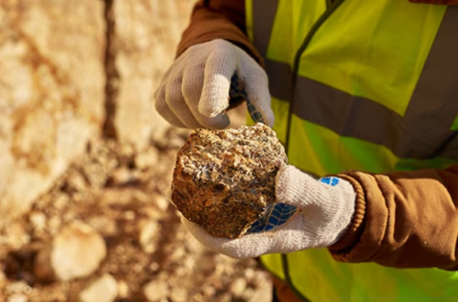 A worker wearing protective gloves and a high-visibility vest examines a rock sample at a mining or geological site.